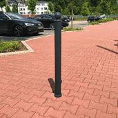 Donkergrijze design afzetpaal Ø76 mm met bolkop, staand op rode klinkers bij een parkeerplaats.