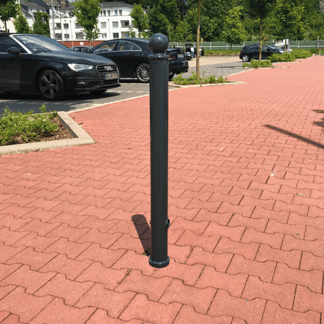 Donkergrijze design afzetpaal Ø76 mm met bolkop, staand op rode klinkers bij een parkeerplaats.