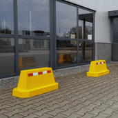 Twee gele betonnen afzetblokken met reflecterende strepen, staand op een bestrate ondergrond.