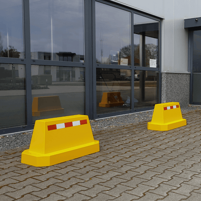 Twee gele betonnen afzetblokken met reflecterende strepen, staand op een bestrate ondergrond.