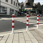 Drie wit-rode afzetpalen van 70x70 mm op een trottoir, met een zebrapad en gebouwen op de achtergrond.