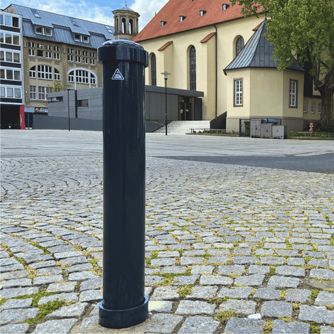 Donkergrijze afzetpaal Ø108 mm met helm kop, staand op een kasseienplein voor een kerk.