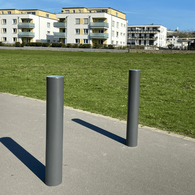Twee donkergrijze, Ø120 mm design afzetpalen met vlakke kop op een pad naast een grasveld en appartementen.