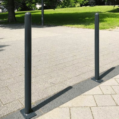 Twee antracietgrijze afzetpalen met puntkop, Ø76 mm, op een tegelpad met groen gras op de achtergrond.