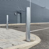 Twee verzinkt stalen beschermpalen Ø89 mm, geplaatst op een parkeerterrein bij een bedrijfsgebouw.
