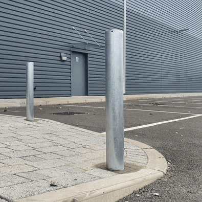 Twee verzinkt stalen beschermpalen Ø89 mm, geplaatst op een parkeerterrein bij een bedrijfsgebouw.
