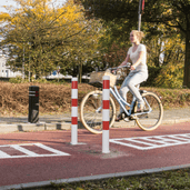 Wit-rode beschermpaal Ø89 mm op een fietspad, met een fietser en een zwarte paal op de achtergrond.
