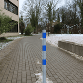 Verzinkt-blauwe beschermpaal Ø60 mm met blauwe reflecterende strepen, staand op een bestraat pad.