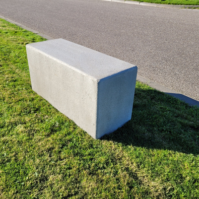 Grijze betonnen poef, rechthoekig, 1000x400x500 mm, op groen gras naast een pad.