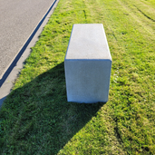 Grijze betonnen poef, 1000x400x500 mm, rechthoekig design, op gras naast een pad.