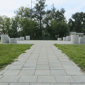 Grijze betonnen poefs Ø700x600 mm, cilindervormig, op een grasveld met bestrating.