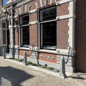 Grijze achthoekige betonnen sierpalen met kettingen voor een bakstenen gevel.
