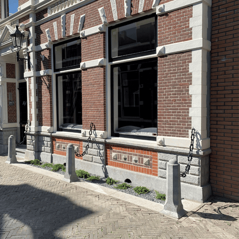 Grijze achthoekige betonnen sierpalen met kettingen voor een bakstenen gevel.