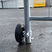 Gegalvaniseerd bouwhekwiel met zwart rubberen band en scharnierbevestiging aan een grijs hekpaal.