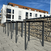 Antraciet fietsbeugel design van plat staal 80x15 mm, langs een gabionmuur bij een modern gebouw.
