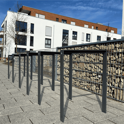 Antraciet fietsbeugel design van plat staal 80x15 mm, langs een gabionmuur bij een modern gebouw.
