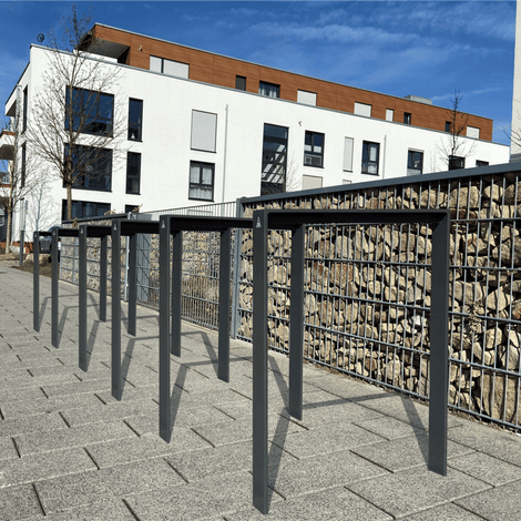 Antraciet fietsbeugel design van plat staal 80x15 mm, langs een gabionmuur bij een modern gebouw.