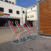 Verzinkt stalen fietsenrek met rode aanleunbeugels, geschikt voor 4 fietsen, buiten een winkelcentrum.