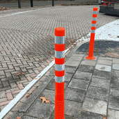 Oranje-witte flexibele paaltjes met reflecterende banden op een stoep.