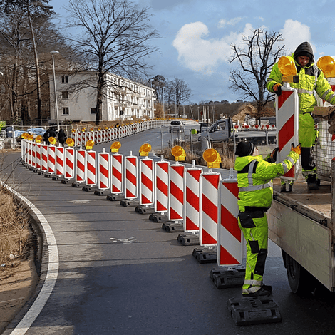 Twee werknemers plaatsen rood-witte geleidebakens met gele lampen langs een weg.