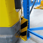 Geel-zwart flexibel hoekprofiel stootbeschermer voor vloermontage, beschermt een blauw magazijnrek tegen een gele heftruck.