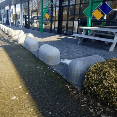 Grijze Jumboblokken van Oud Hollands stampbeton, halfrond, langs een trottoir met struiken en een gebouw.