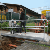Zilveren aluminium loopbrug met veiligheidsrailing, over een bouwput, met een vrouw en kinderwagen.
