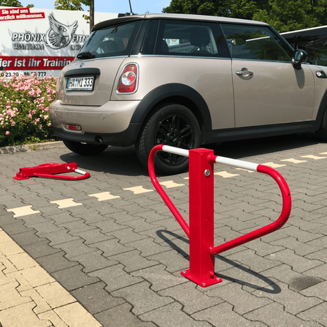 Rood-witte parkeerbeugel met U-vormige beugel, klaar voor betonverankering, naast een geparkeerde auto.