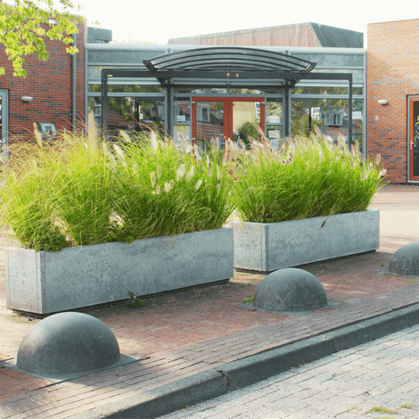 Antraciet betonnen parkeerbol Ø500 mm op voet, naast betonnen plantenbakken met groen gras.