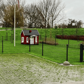 Grijze betonnen parkeerbol op voet, Ø350 mm, naast een groen gazon en een rood huisje.