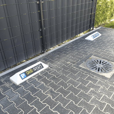 Witte stootbanden met blauw-geel "P DIE-SIGN" logo op een grijze klinkerparkeerplaats.