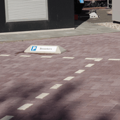 Grijze betonnen stootband met gepersonaliseerd "P Bezoekers" parkeerbord op een klinkerparkeerplaats.