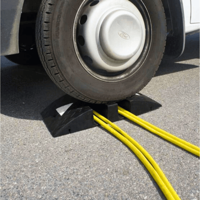 Zwarte rubberen slangenbrug met 2 kanalen (95x90mm) onder een voertuigwiel, beschermt gele kabels.