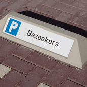 Grijze betonnen stootband met "Bezoekers" tekst en blauw P-symbool op een parkeerplaats.