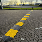 Grijze en gele verkeersdrempel van 3 cm hoog op een parkeerplaats, met vrachtwagens op de achtergrond.