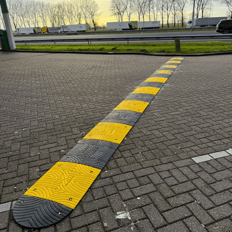 Grijze en gele verkeersdrempel van 3 cm hoog op een parkeerplaats, met vrachtwagens op de achtergrond.