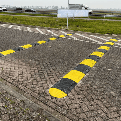 Zwarte en gele rubberen verkeersdrempel van 5 cm hoog op een parkeerplaats, met een snelweg op de achtergrond.