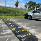 Zwarte rubberen verkeersdrempel van 7 cm met gele reflecterende markeringen op asfalt, naast een zilveren bestelwagen.