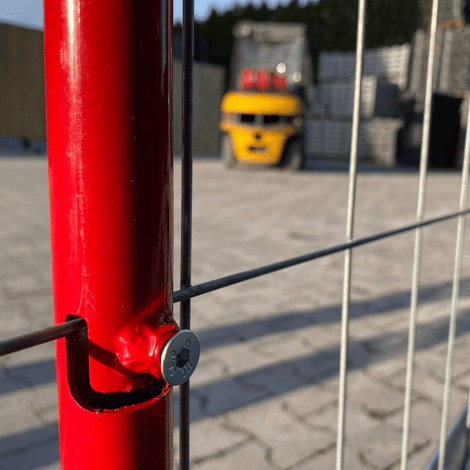 Rood-witte waarschuwingsbalk voor bouwhekken, bevestigd met metalen steunen aan een rood hek.