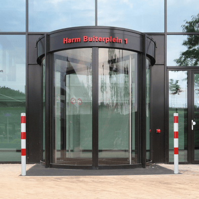 Twee wit-rode parkeerpalen Ø60 mm voor verankering, geplaatst voor een modern gebouw met draaideur.