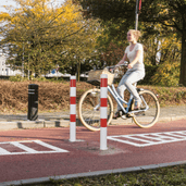 Wit-rode parkeerpaal met betonvoet op fietspad, Ø60-102 mm, naast fietsende vrouw.